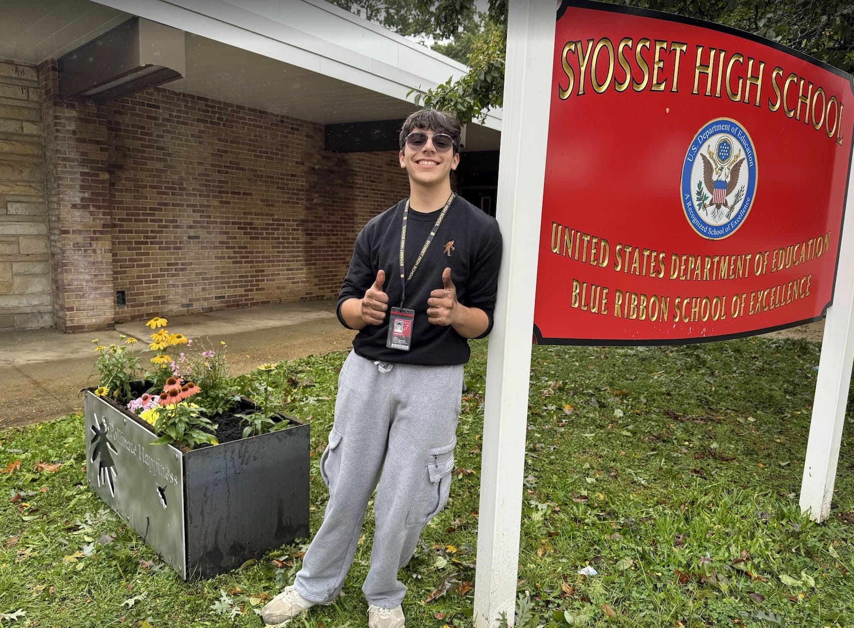 Aidan Ellis giving a thumbs up beside the new Corten steel pollinator planter with its laser-cut aluminum plaque, next to the red Syosset High School sign